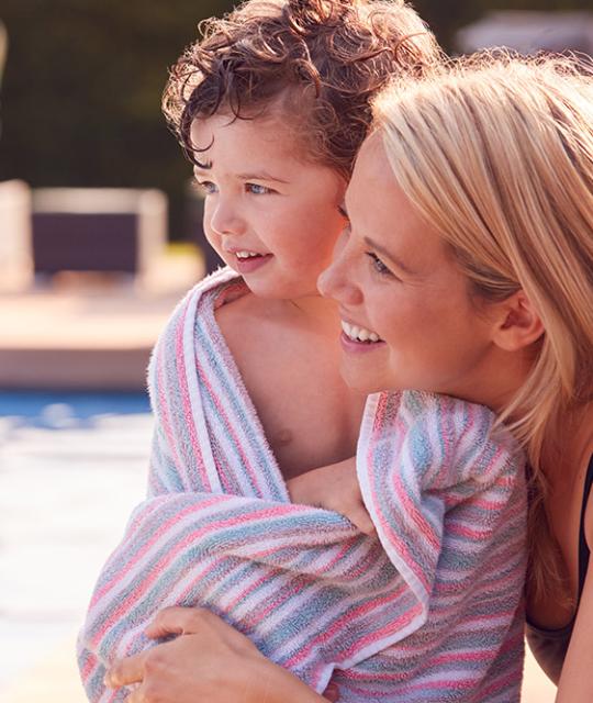 Madre e figlio sorridenti vicino a una piscina, avvolti in un asciugamano colorato.