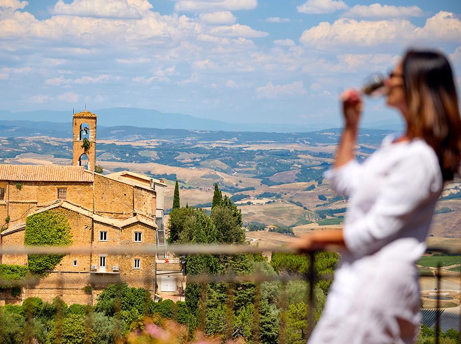 Donna beve vino con vista su paesaggio toscano e chiesa antica.