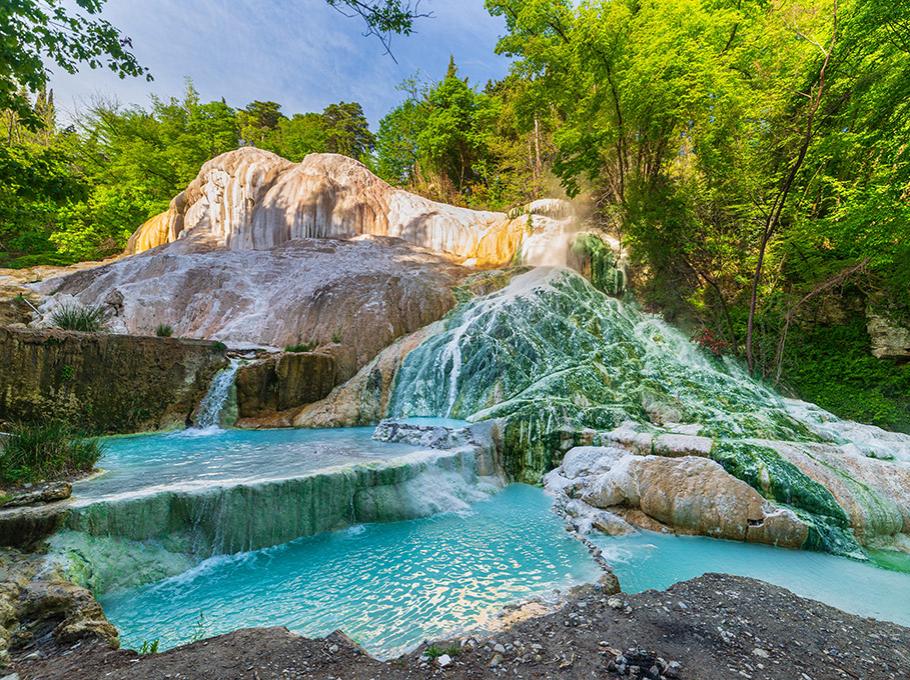 Cascata termale con acqua azzurra e rocce calcaree circondate da vegetazione lussureggiante.