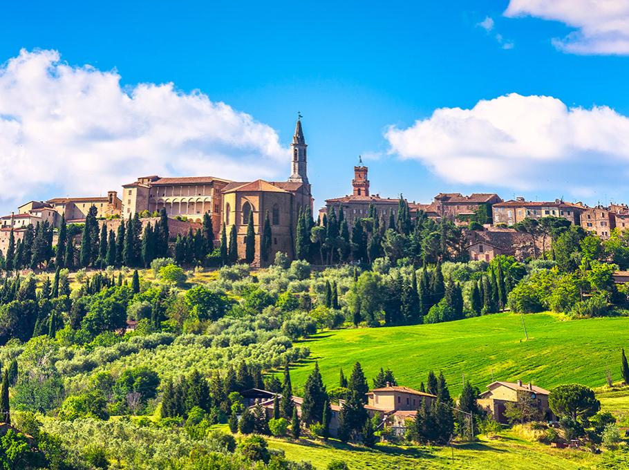 Paesaggio collinare toscano con cipressi, edifici storici e cielo azzurro.