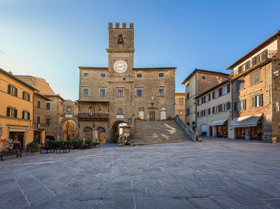 Piazza storica con palazzo antico e orologio, circondata da edifici tradizionali.